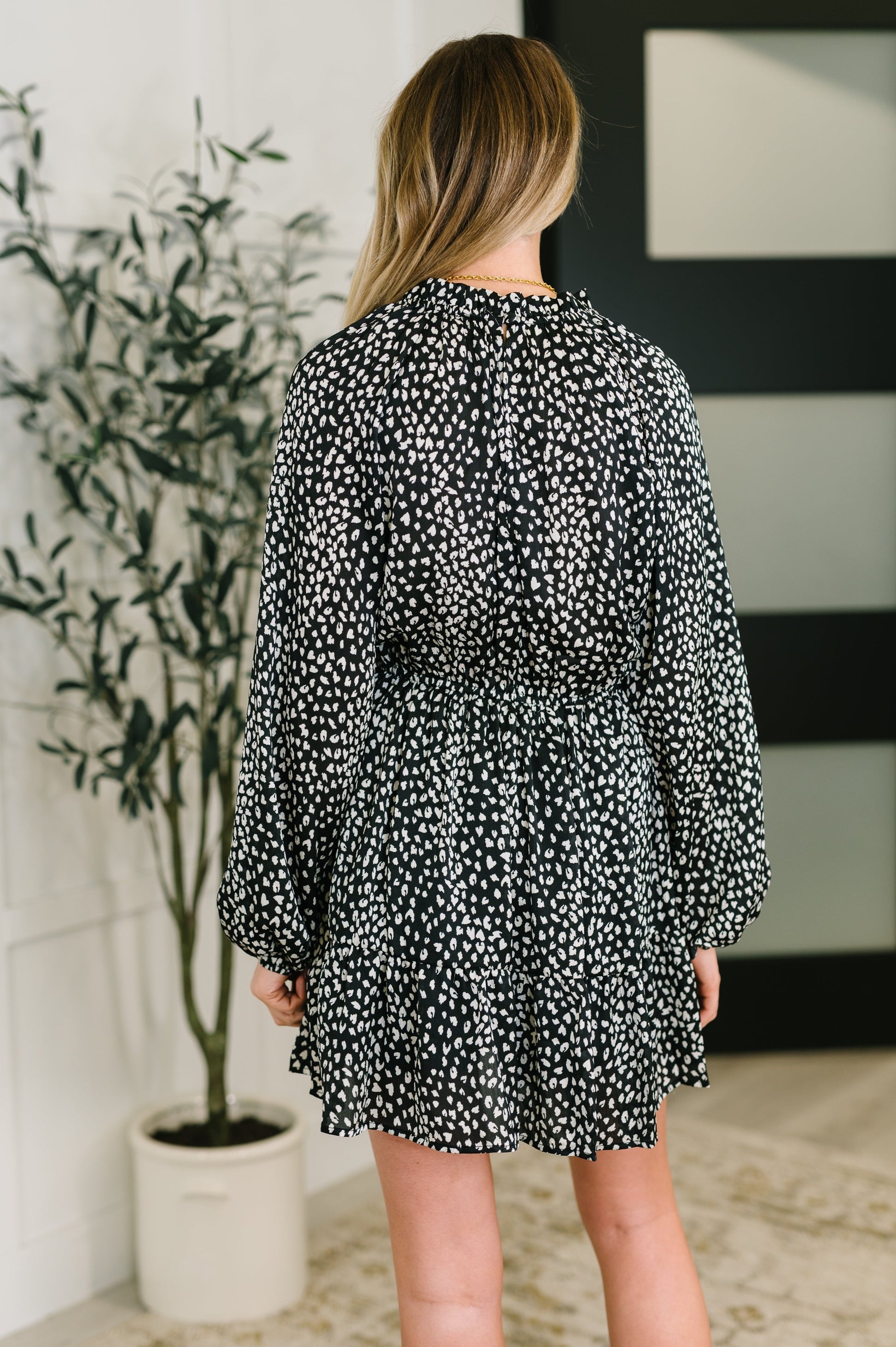 Woman wearing a black and white patterned dress in a room with a plant and wall art.