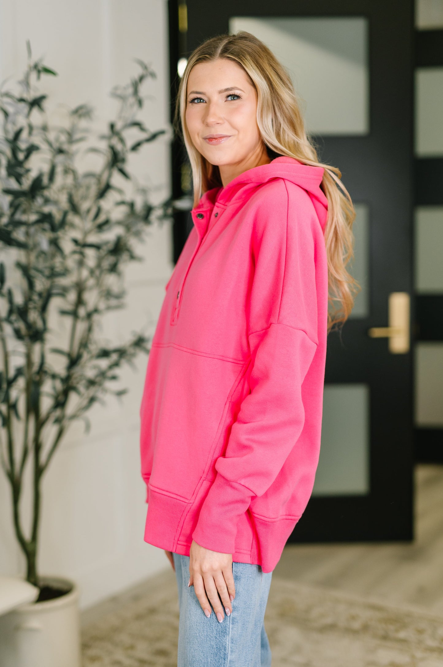 Woman wearing a bright pink hoodie standing indoors next to a plant.