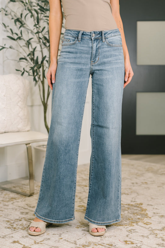 Person wearing light blue flared jeans in a room with a plant and bench.