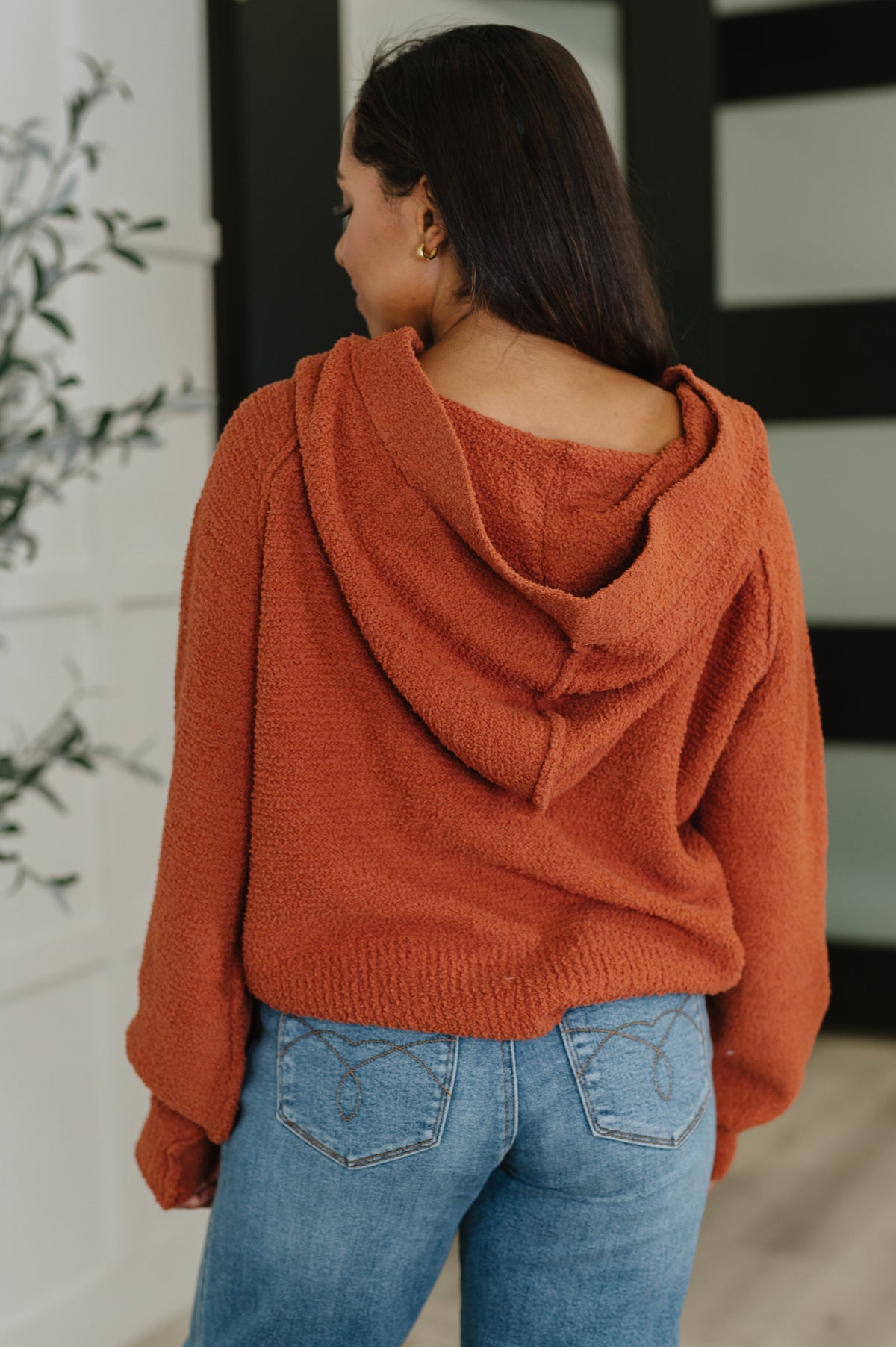 A woman shown from the back wearing a fuzzy, oversized rust-orange hooded sweater and light blue jeans in an indoor setting.