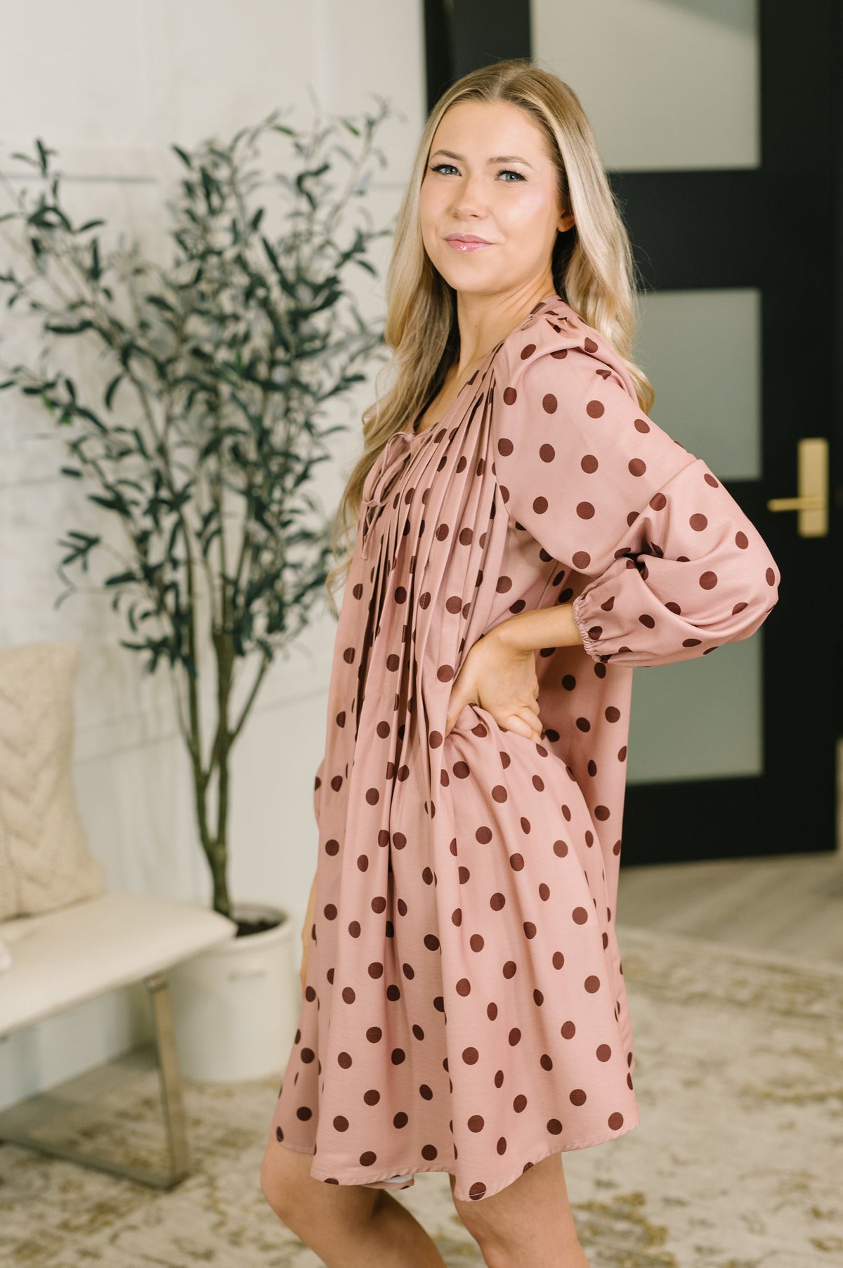A woman modeling a dusty pink long-sleeve dress with dark brown polka dots, standing in a brightly lit indoor setting with a potted plant and a bench in the background.