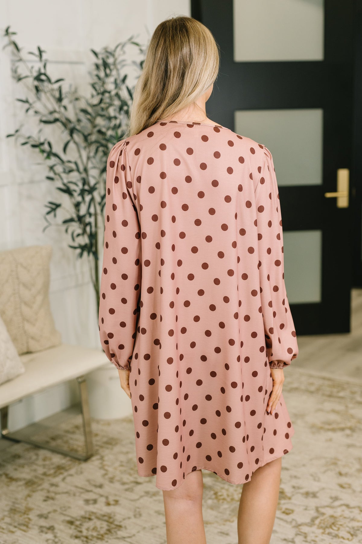 A woman viewed from behind wearing a pink long-sleeve tunic dress with dark brown polka dots, standing in a modern indoor setting with a bench and plant in the background.