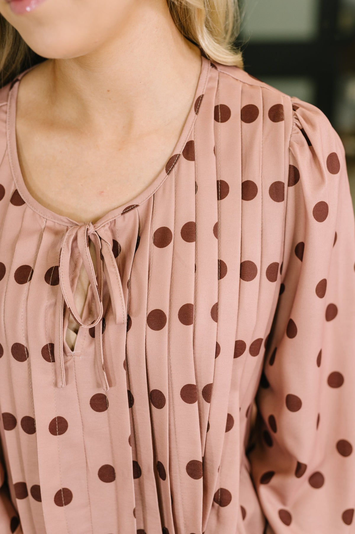 Close-up of a woman wearing a dusty pink long-sleeve blouse with dark brown polka dots and a tie neckline.