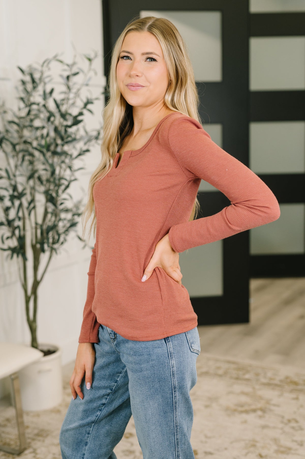 A woman modeling a long-sleeve scoop-neck knit top in a dusty rose color, paired with blue jeans, standing in a modern indoor setting with a plant and a black-framed glass door in the background.