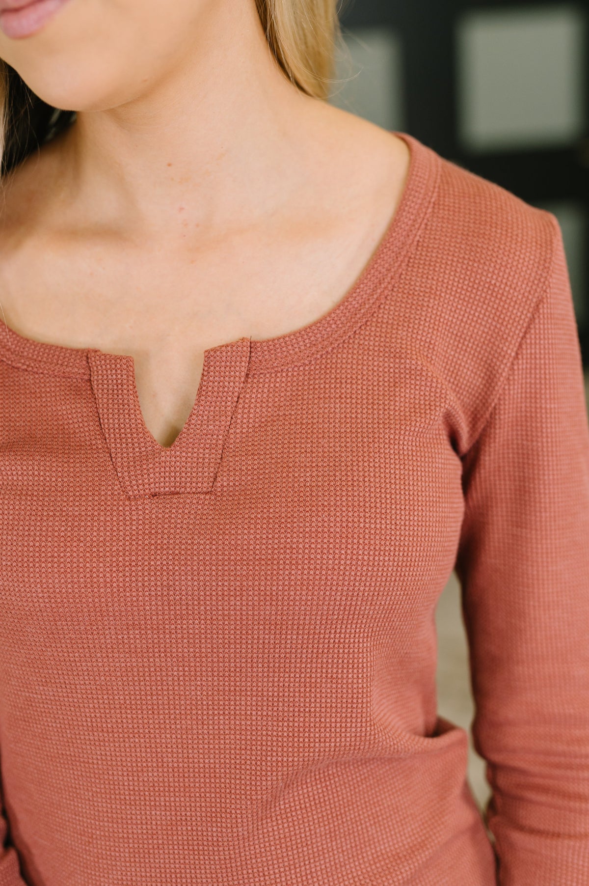 Close-up of a woman wearing a textured, long-sleeve waffle-knit top in a muted terracotta or rust color, featuring a split V-neckline.