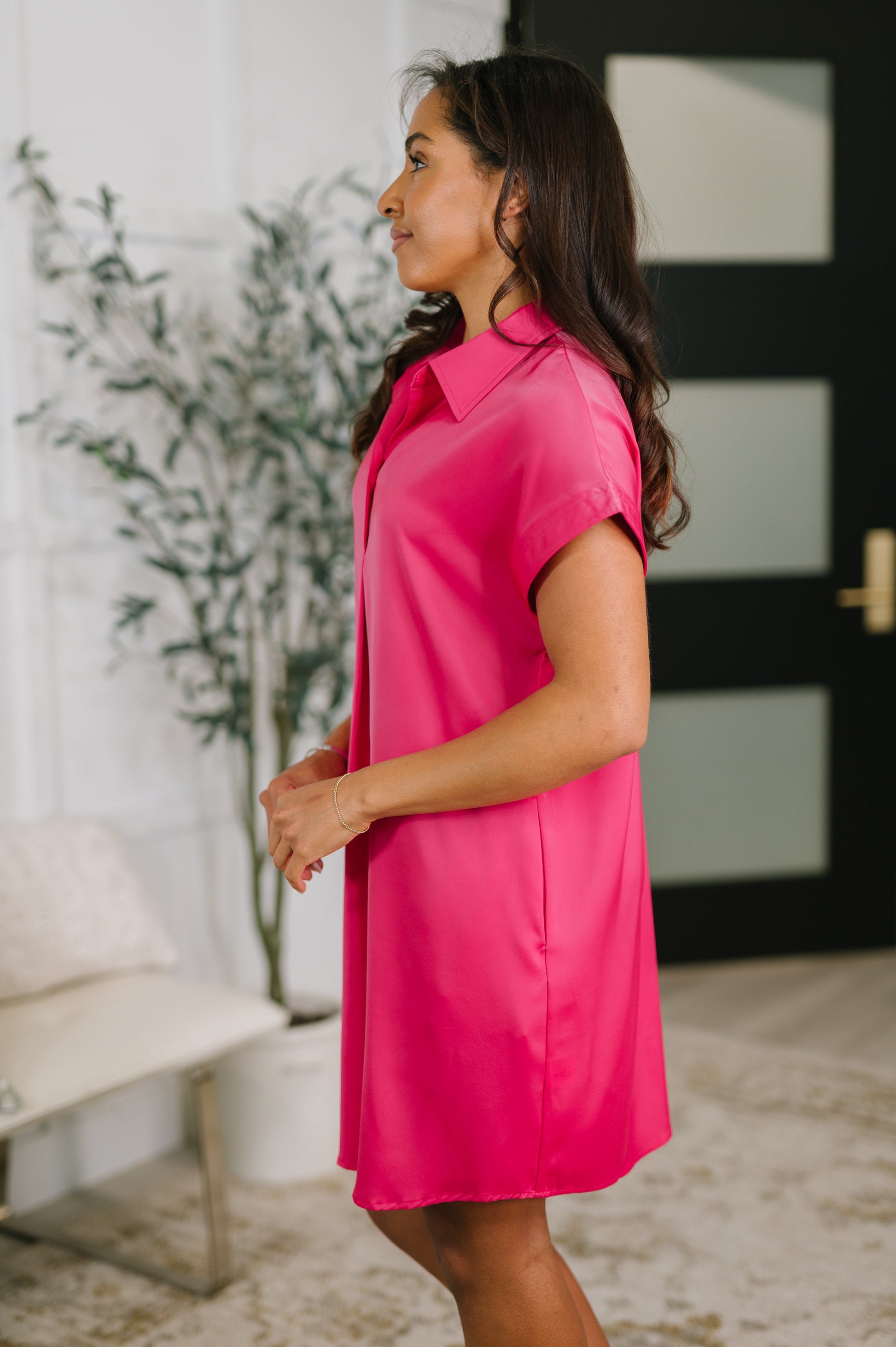 Woman wearing a bright pink dress standing indoors with a decorative plant and door in the background.