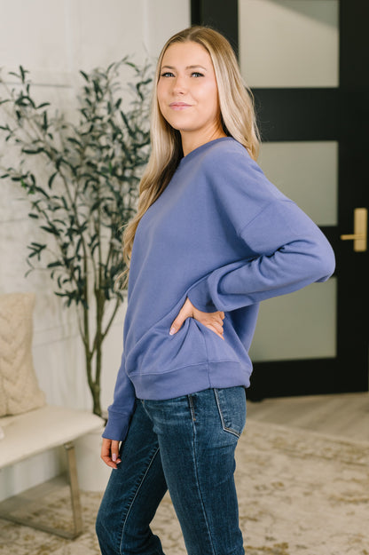 A woman wearing a dusty purple long-sleeve sweatshirt and dark blue jeans, standing in a modern indoor room with a plant and a bench in the background.