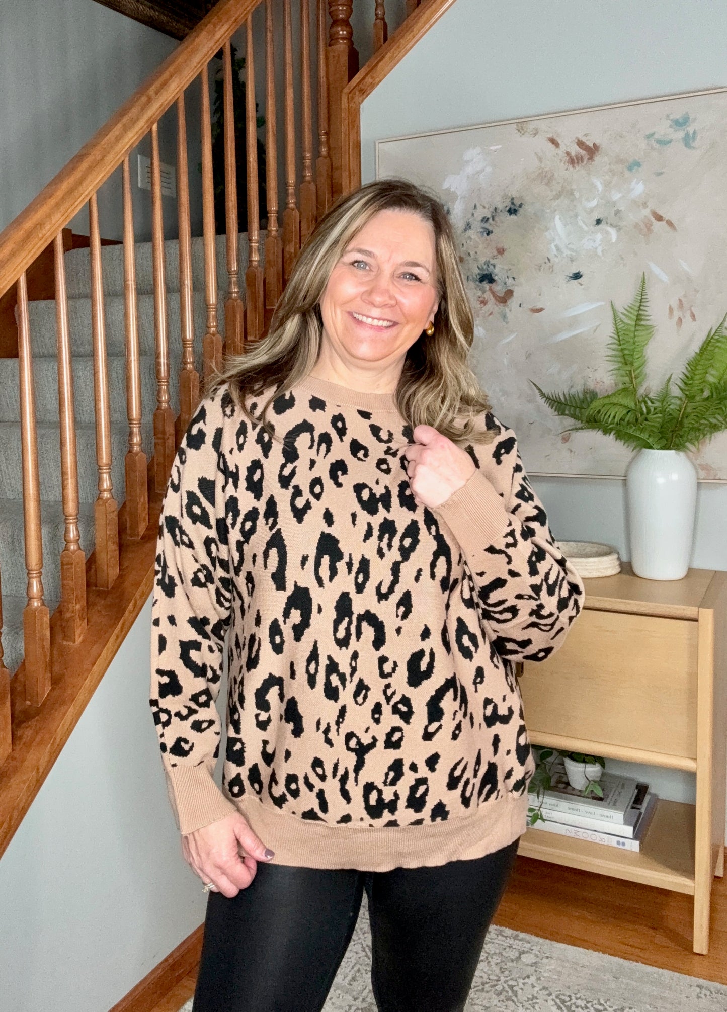 Woman wearing a leopard print sweater in a home setting with a staircase and decor.