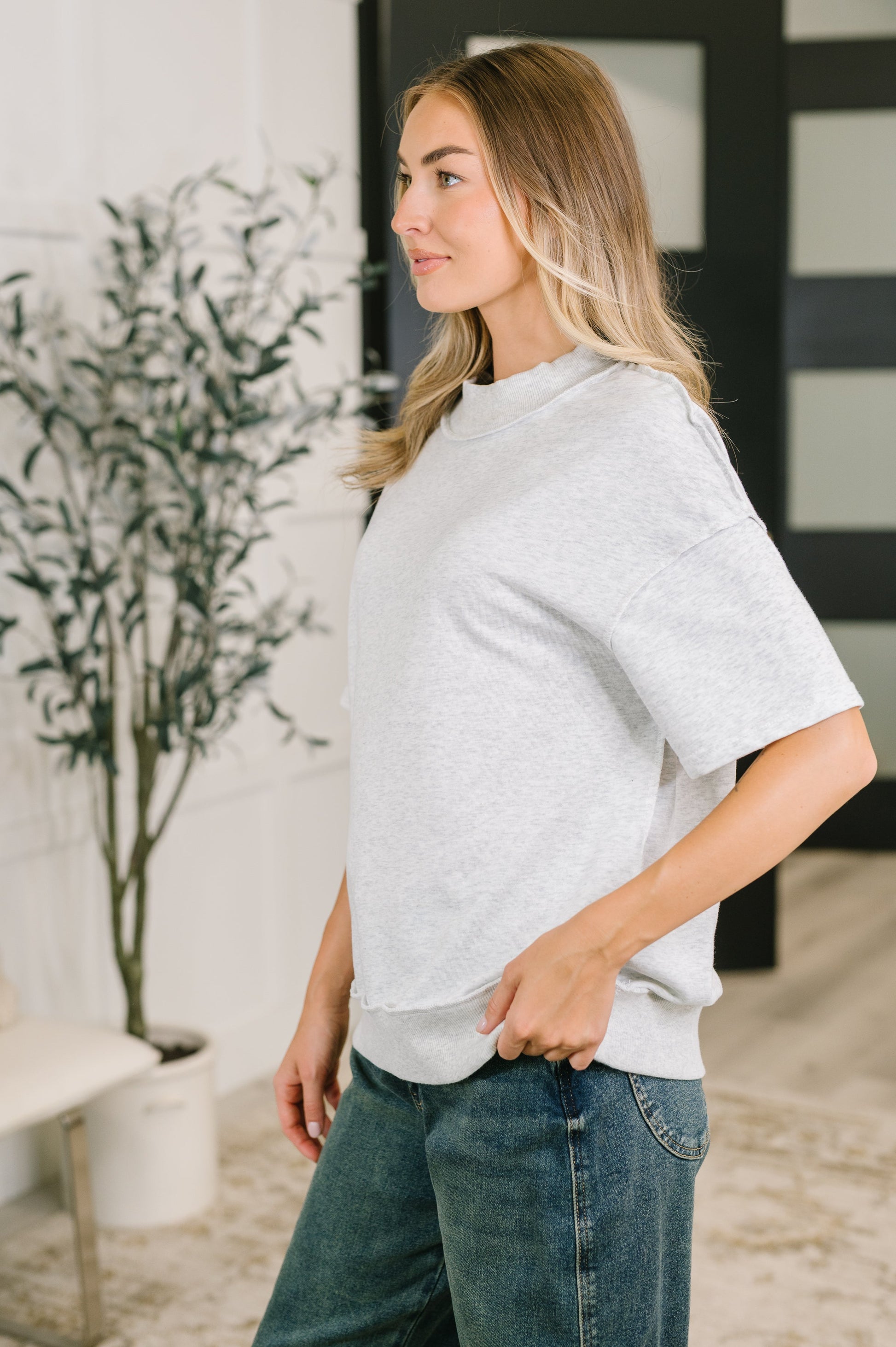 A woman wearing a light heather gray oversized short-sleeve crewneck sweatshirt and blue jeans, standing in a brightly lit indoor setting.