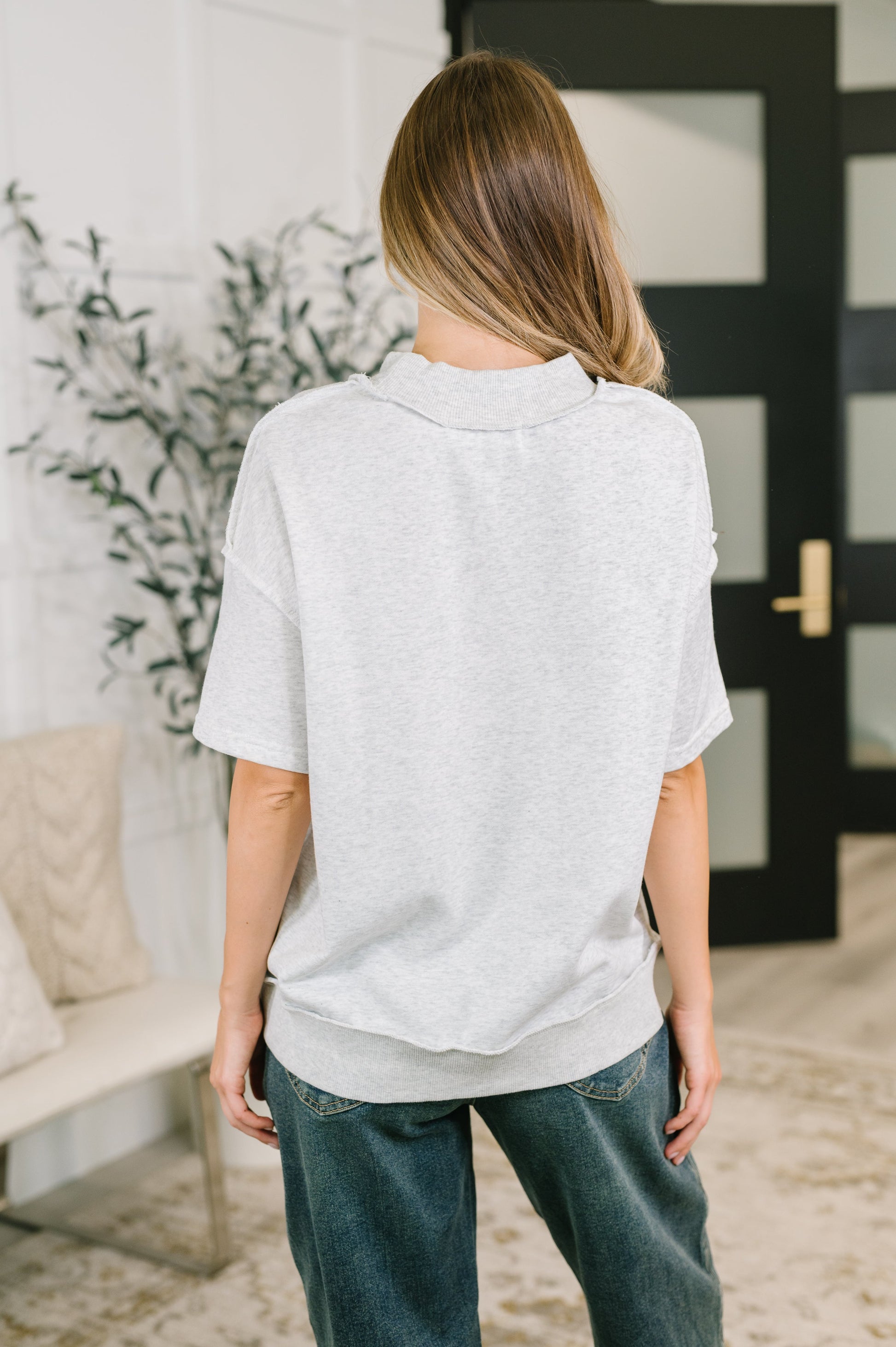 A woman viewed from behind wearing an oversized light gray short-sleeve top and blue jeans in a bright indoor room with a black door and plant in the background.
