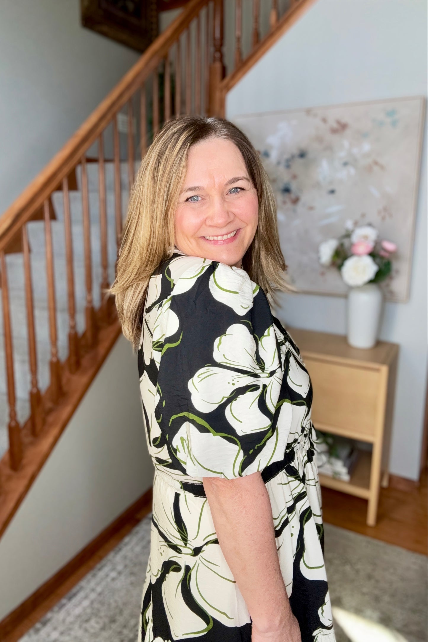 Woman wearing a black and white floral dress standing in a home interior with a staircase and decorative items.