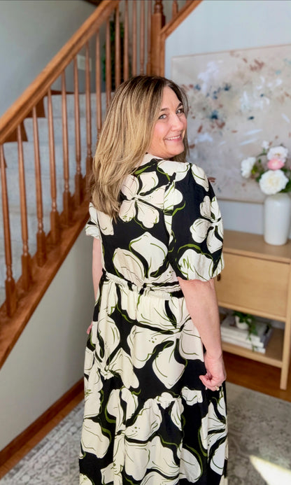 Woman wearing a black floral dress standing in a home setting with a staircase and decor.