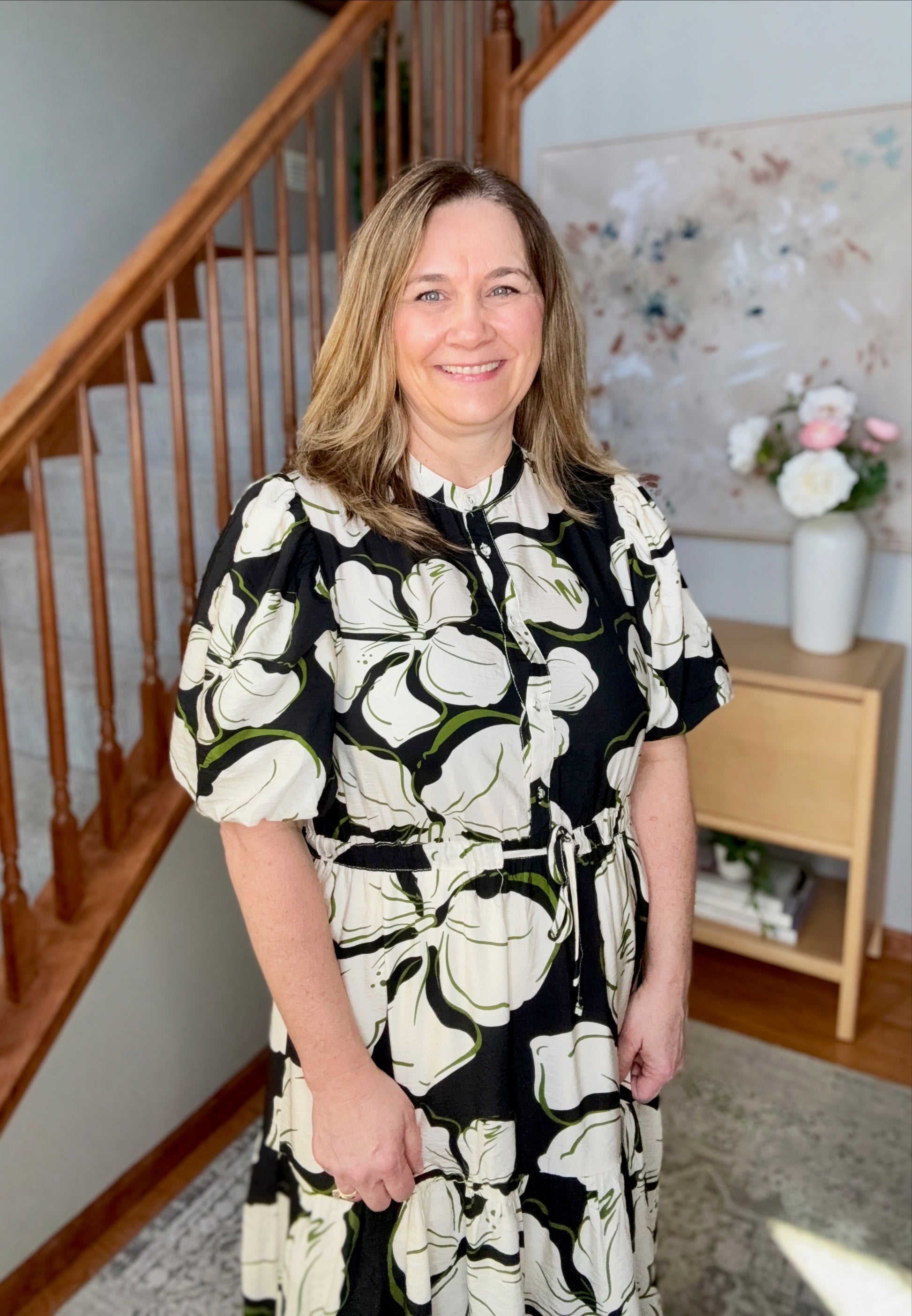 Woman in a floral dress standing in a home interior with a staircase and decor.
