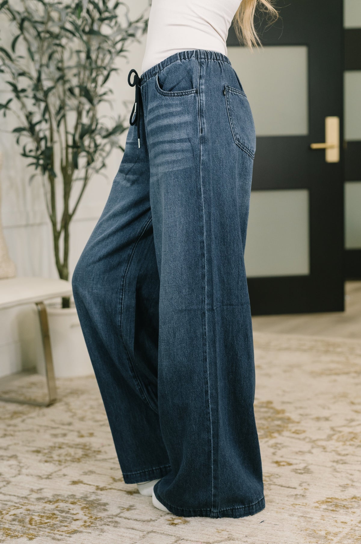 A person wearing dark blue wide-leg denim jeans with a drawstring waistband, standing in an indoor room with a patterned rug, potted plant, and dark door in the background.