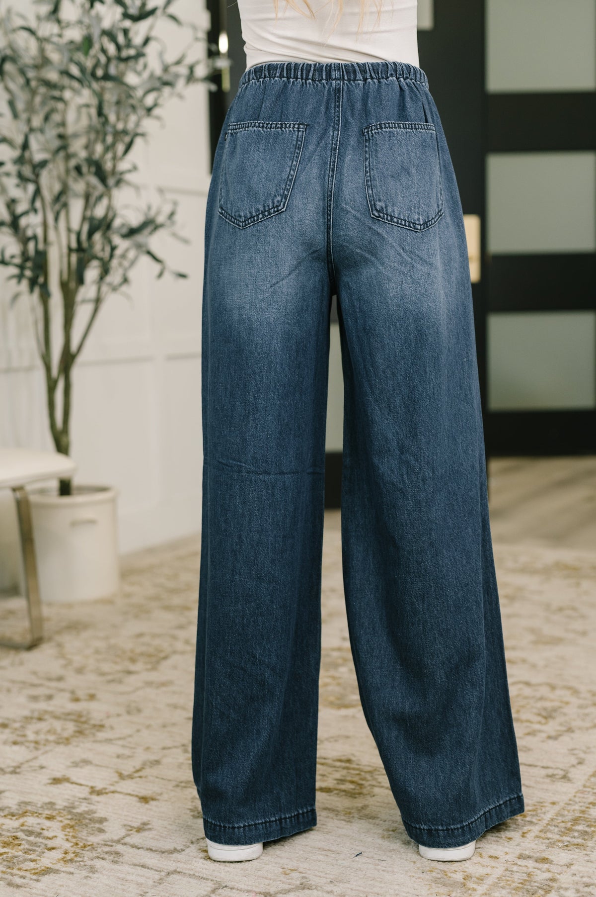 A person shown from the back wearing wide-leg blue denim jeans with an elastic waistband, standing indoors on a patterned rug.