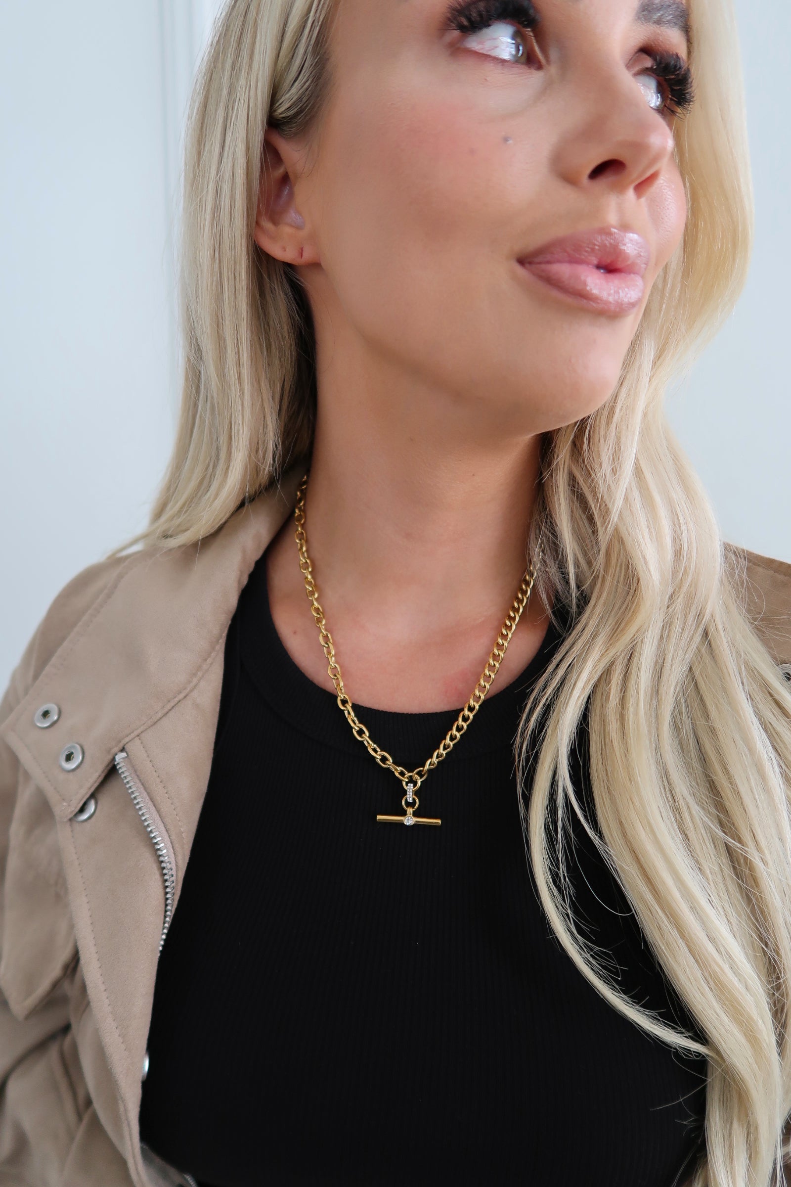 Woman wearing a gold necklace with a black top and beige jacket.