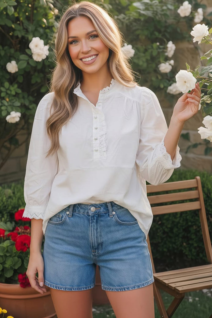 Woman wearing a white blouse and blue shorts standing in a garden with flowers and plants.