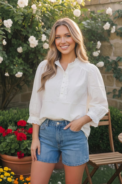 Woman in a white blouse and denim shorts standing in a garden with flowers and a stone wall.