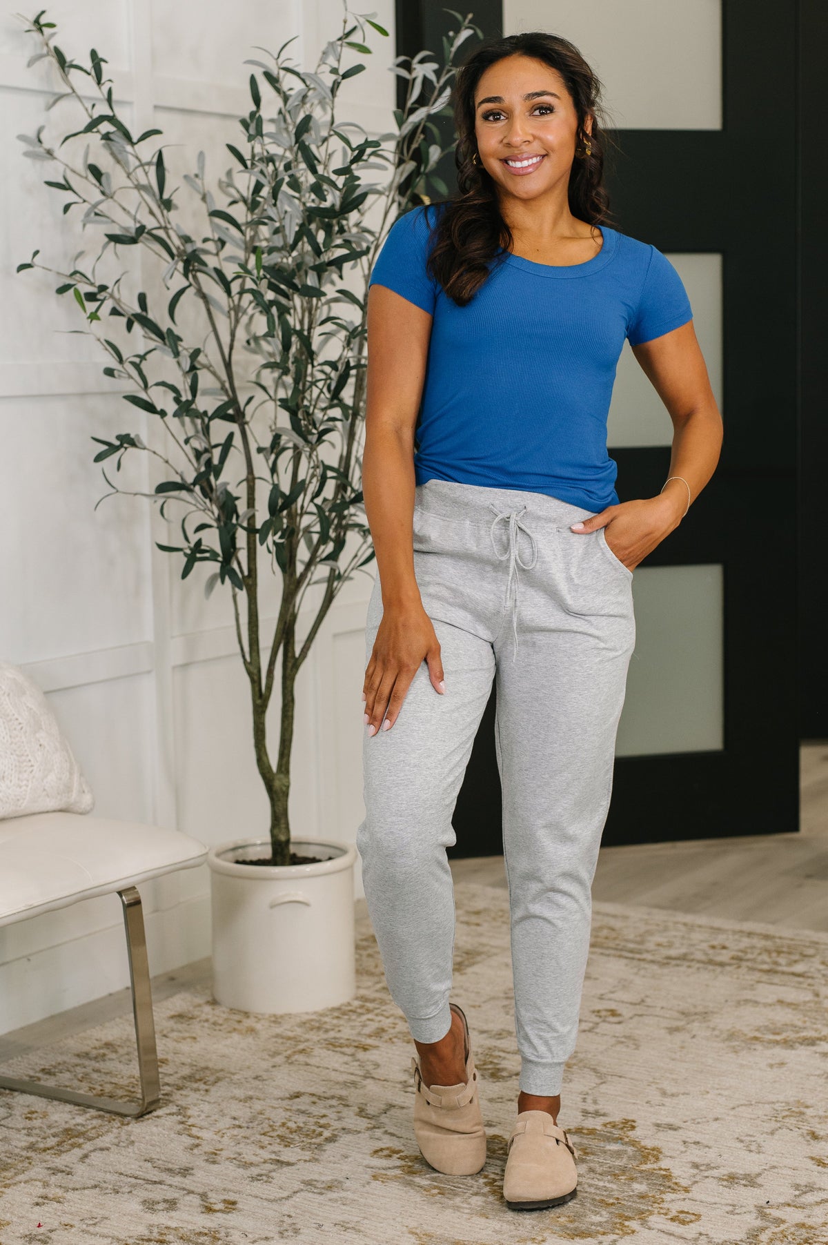 Woman wearing a blue shirt and gray pants standing in a room with a plant and furniture.