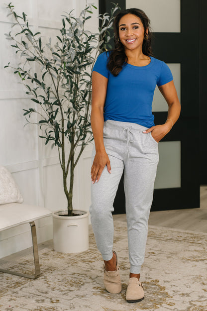 Woman wearing a blue shirt and gray pants standing in a room with a plant and furniture.
