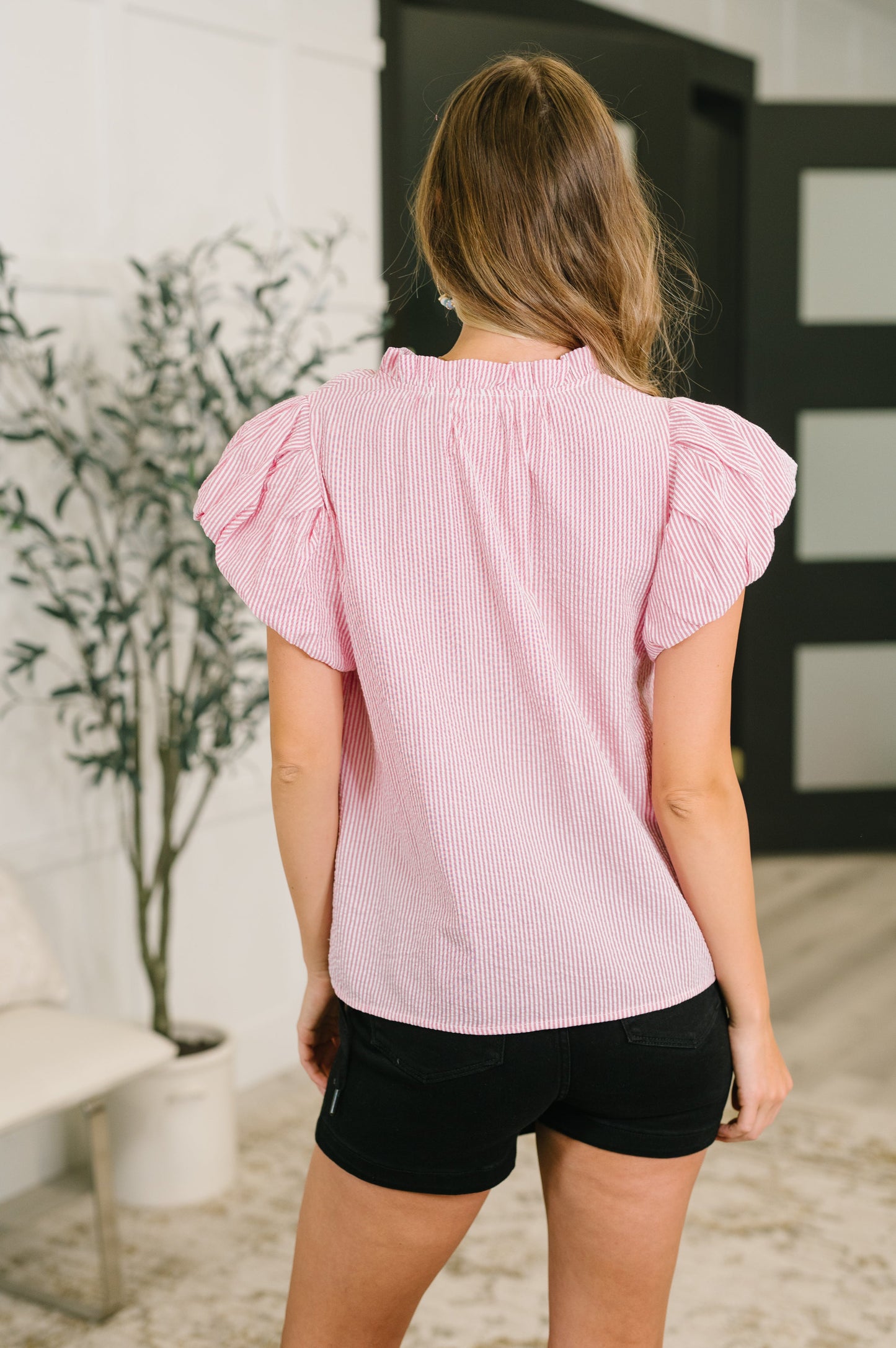 A woman viewed from the back wearing a light pink and white vertically striped short-sleeve blouse with ruffled shoulders, paired with black shorts, standing in an indoor setting.