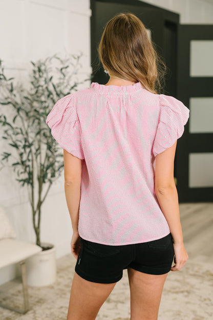 A woman viewed from the back wearing a light pink and white vertically striped short-sleeve blouse with ruffled shoulders, paired with black shorts, standing in an indoor setting.
