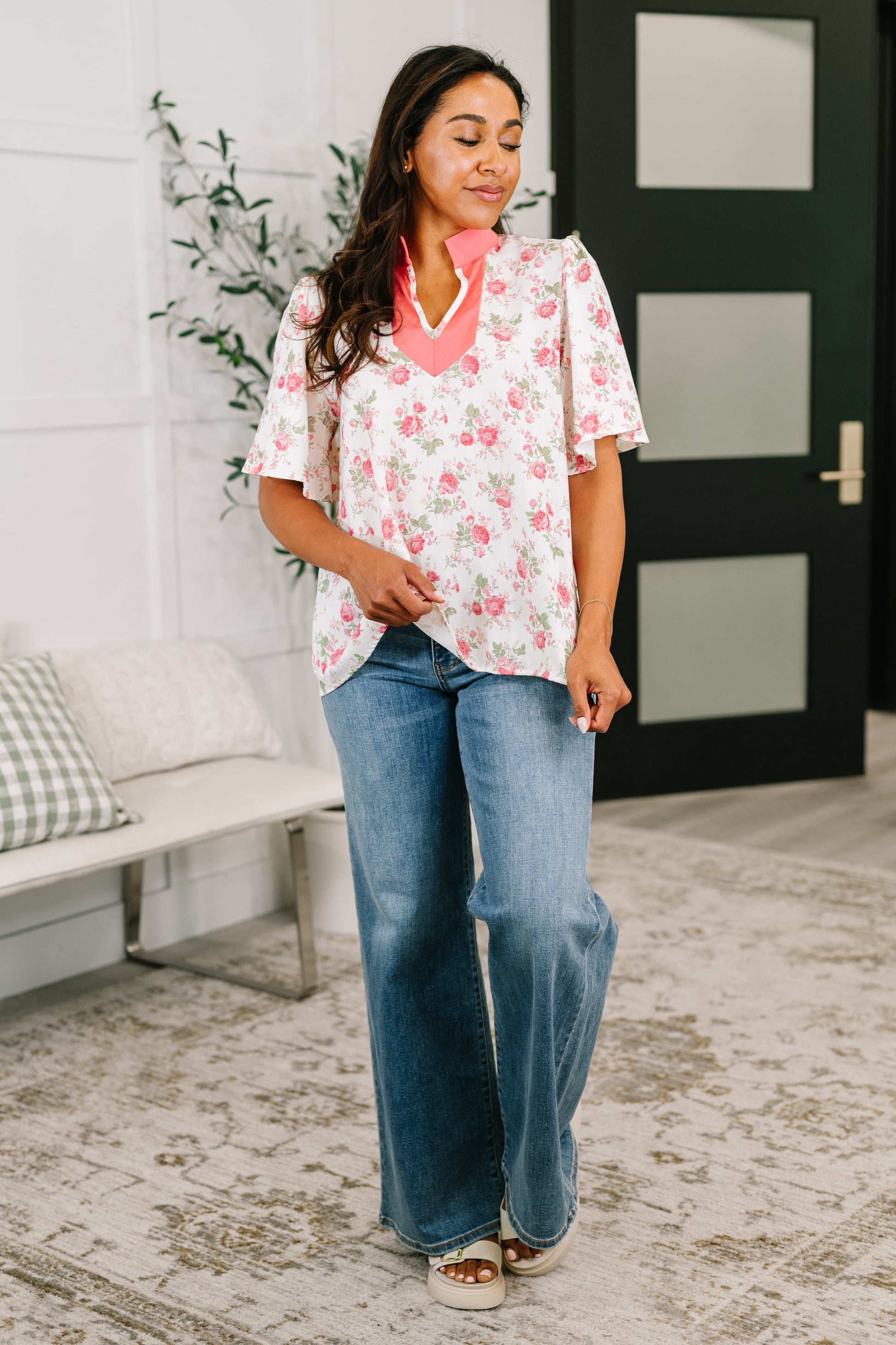 Woman wearing a floral blouse and jeans standing in a room with a white wall and door.