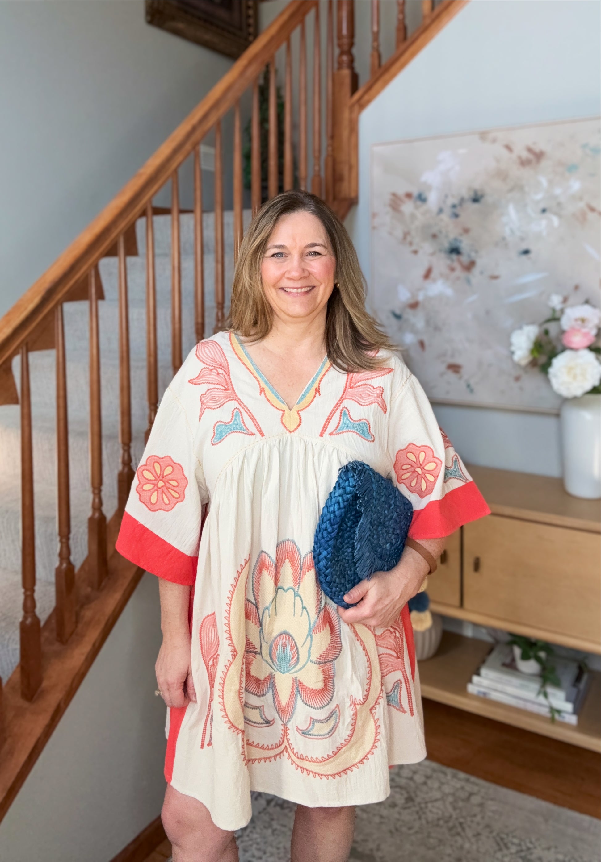 Woman in a patterned dress holding a clutch, standing in a home setting with a staircase and decor.