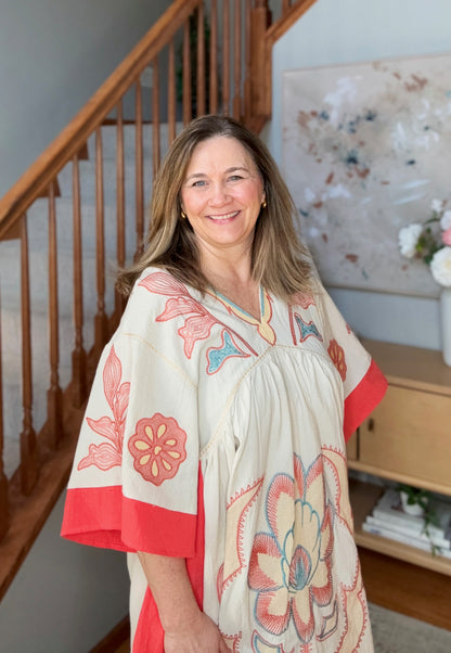 Woman wearing a floral dress standing in a home setting with a staircase and artwork in the background.