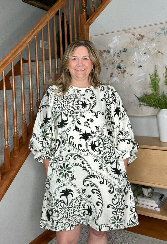 Woman wearing a black and white patterned dress standing in a home setting with a staircase and decor.