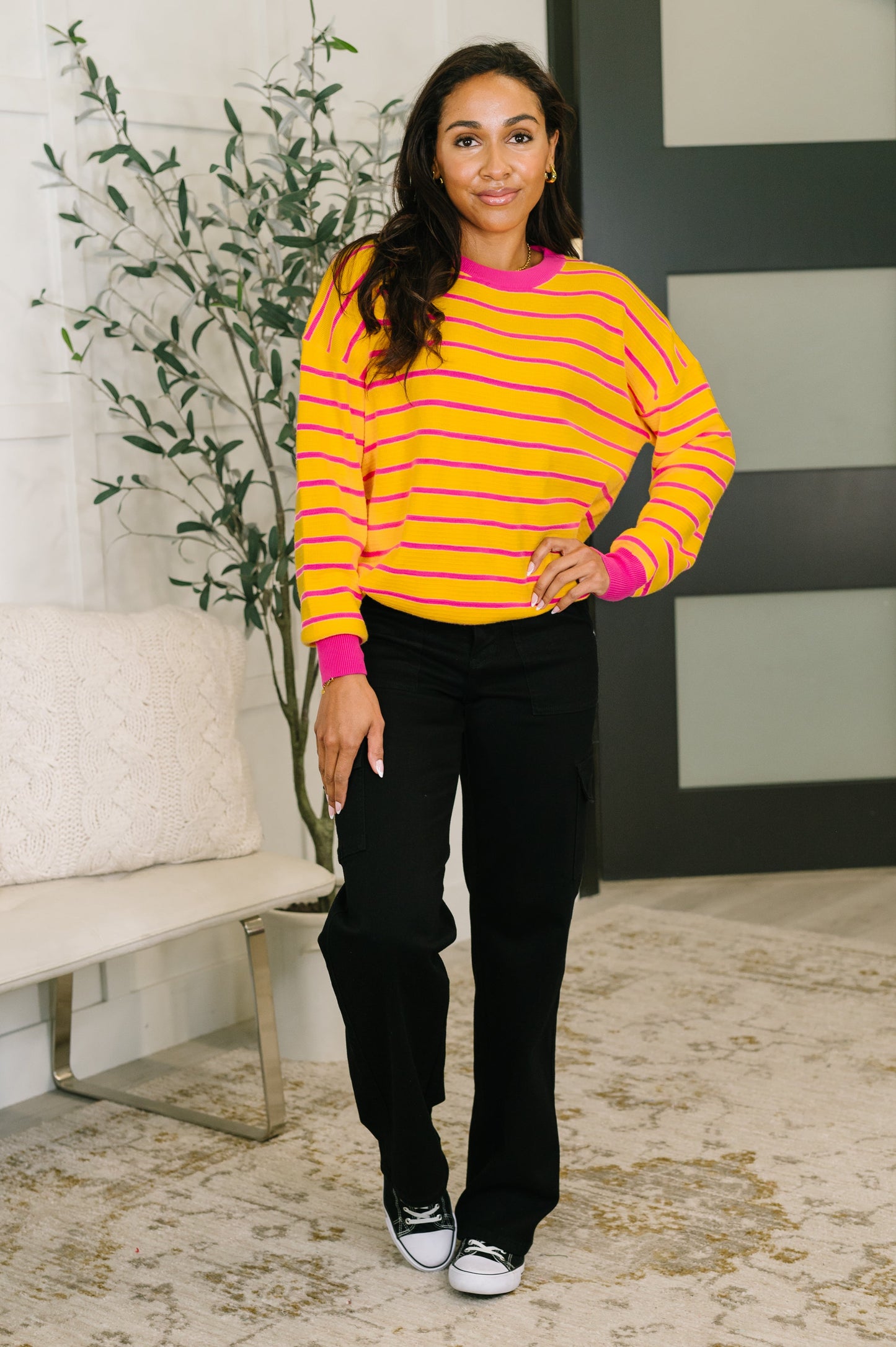 Woman wearing a yellow and pink striped sweater with black pants in an indoor setting.