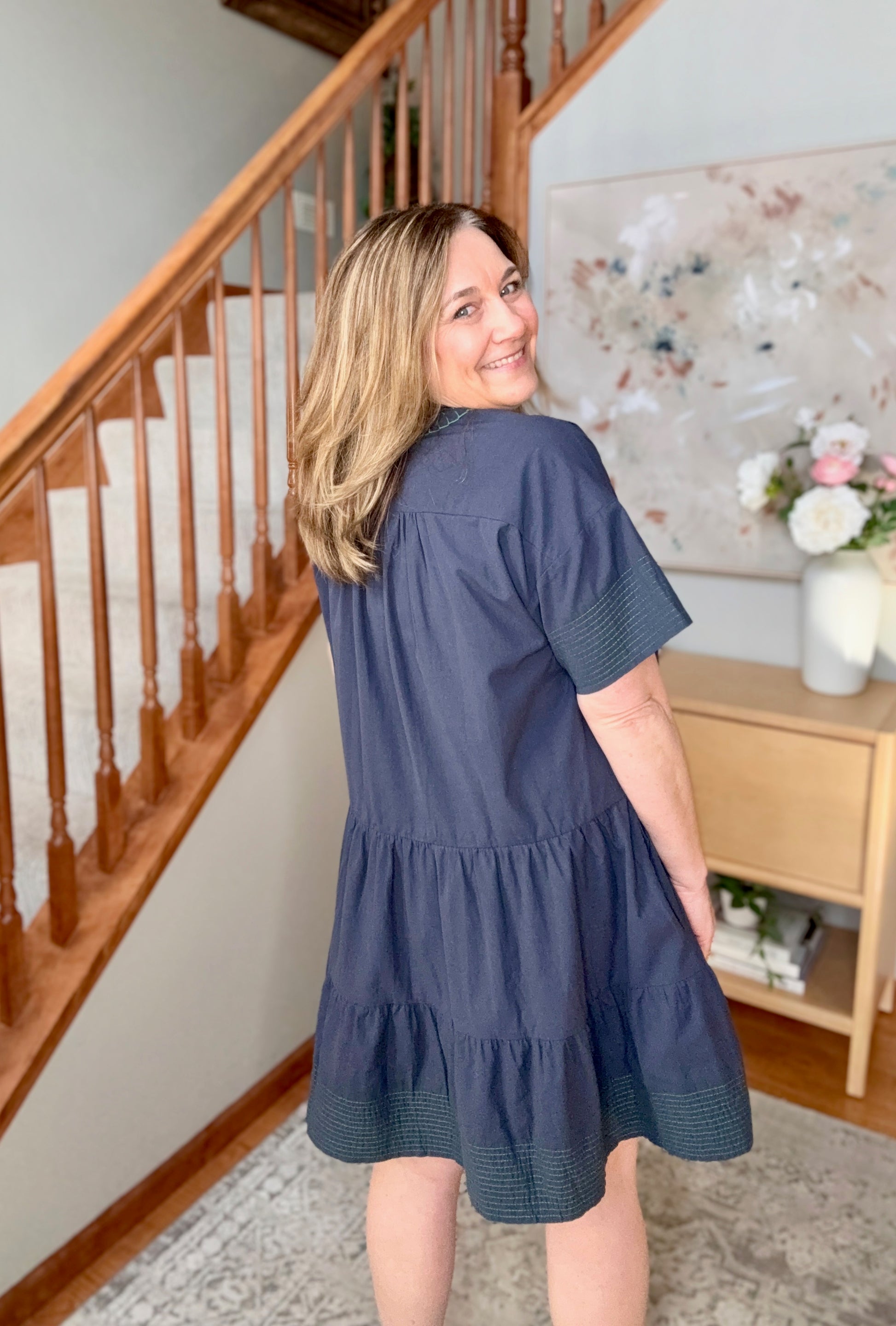 Woman in a blue dress standing in a home setting with a staircase and decorative items.
