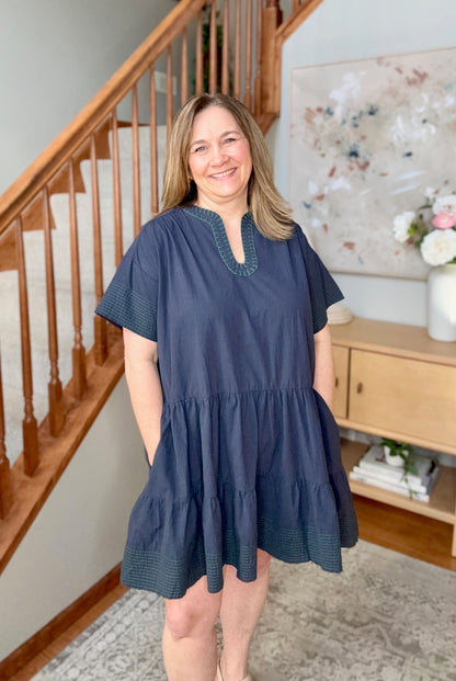 Woman wearing a blue dress standing in a home setting with a staircase and decor.