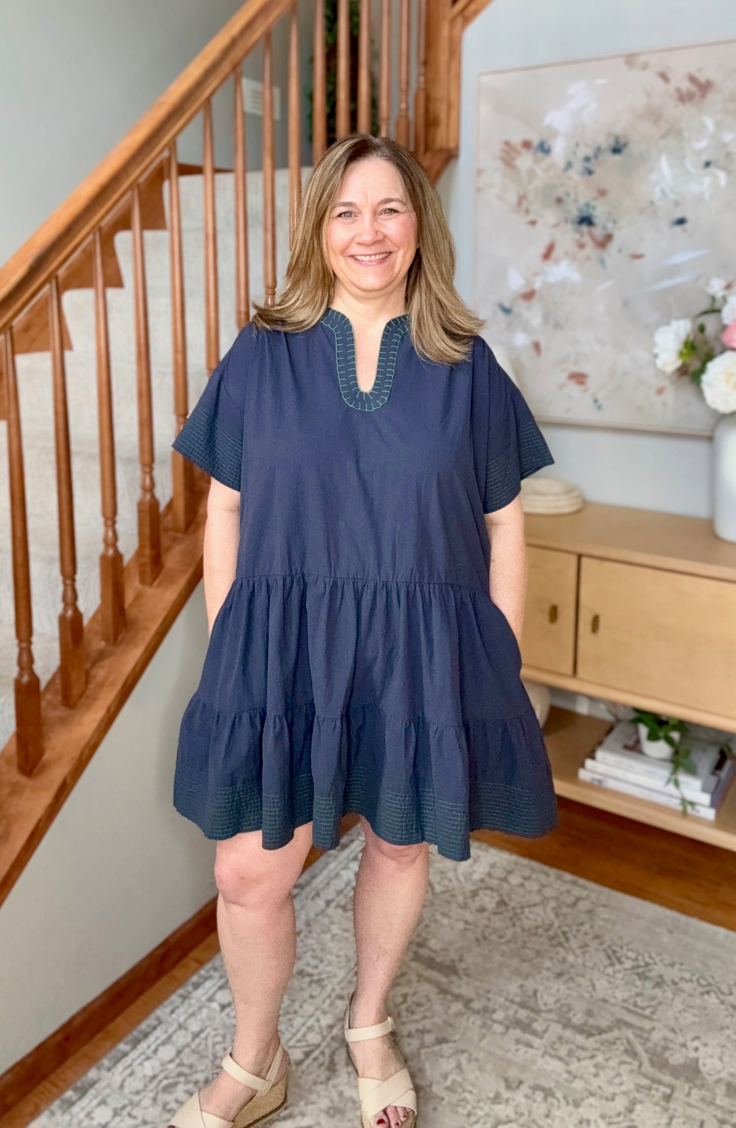 Woman wearing a blue dress standing in a home interior with wooden staircase and decor.