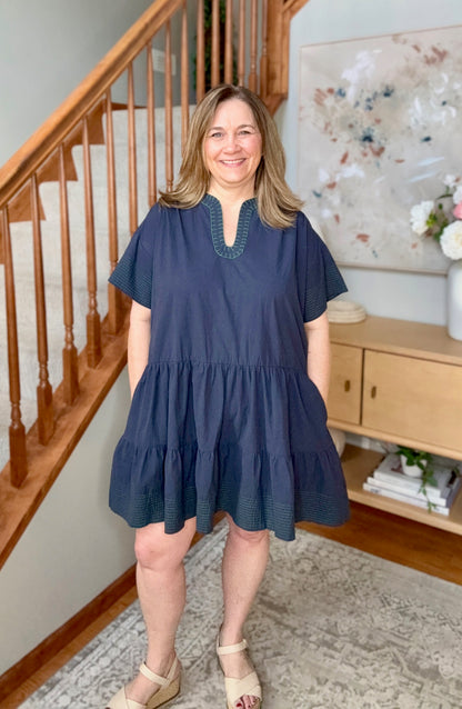 Woman wearing a blue dress standing in a home interior with wooden staircase and decor.