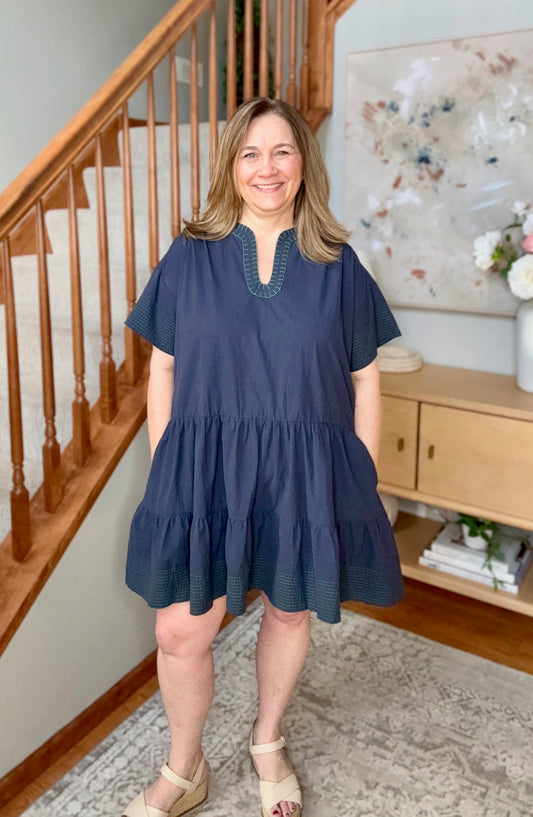 Woman wearing a blue dress standing in a home interior with wooden staircase and decor.