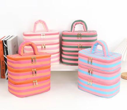 Four striped zippered cosmetic or travel cases with top handles are arranged on a white shelf and surface. The cases come in pink-and-white, orange-and-pink, and pink-and-blue color combinations, with gold-tone zipper hardware.