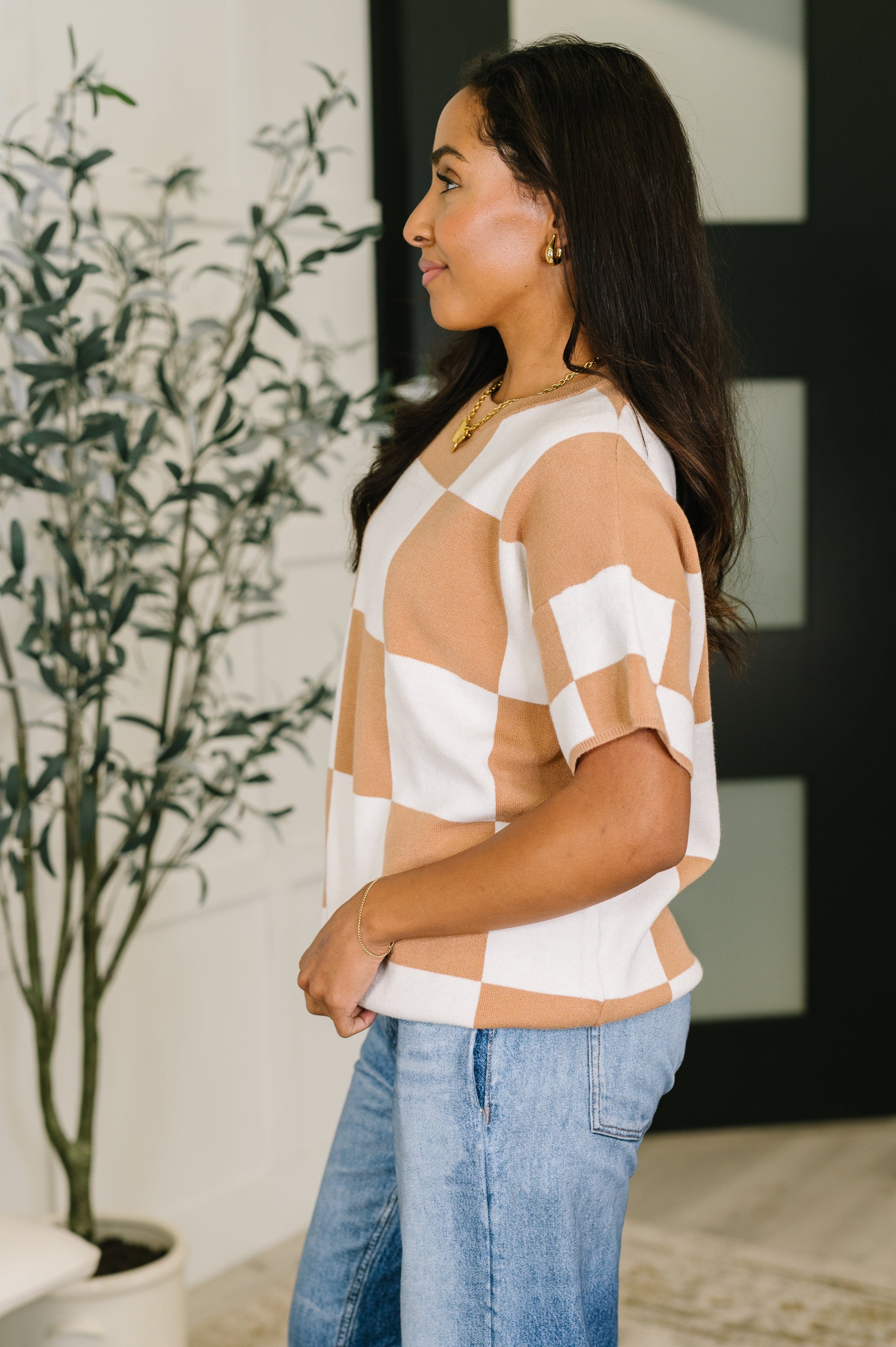 Woman wearing a checkered top and jeans standing indoors with a plant in the background