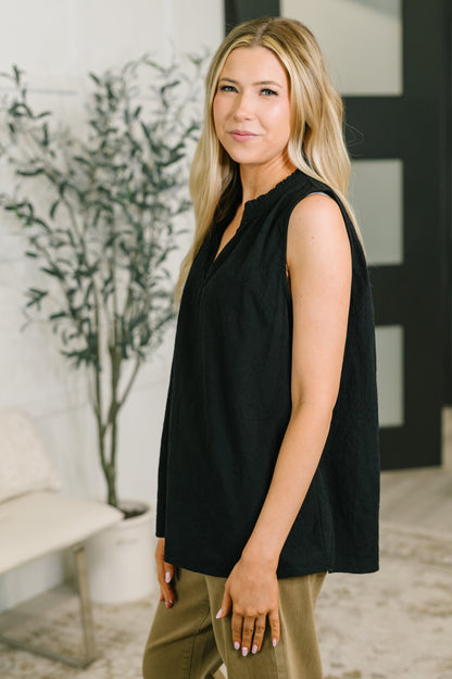 A woman modeling a sleeveless black top with a split neckline, paired with olive green pants, standing in a modern indoor setting with a potted plant and light-colored bench in the background.