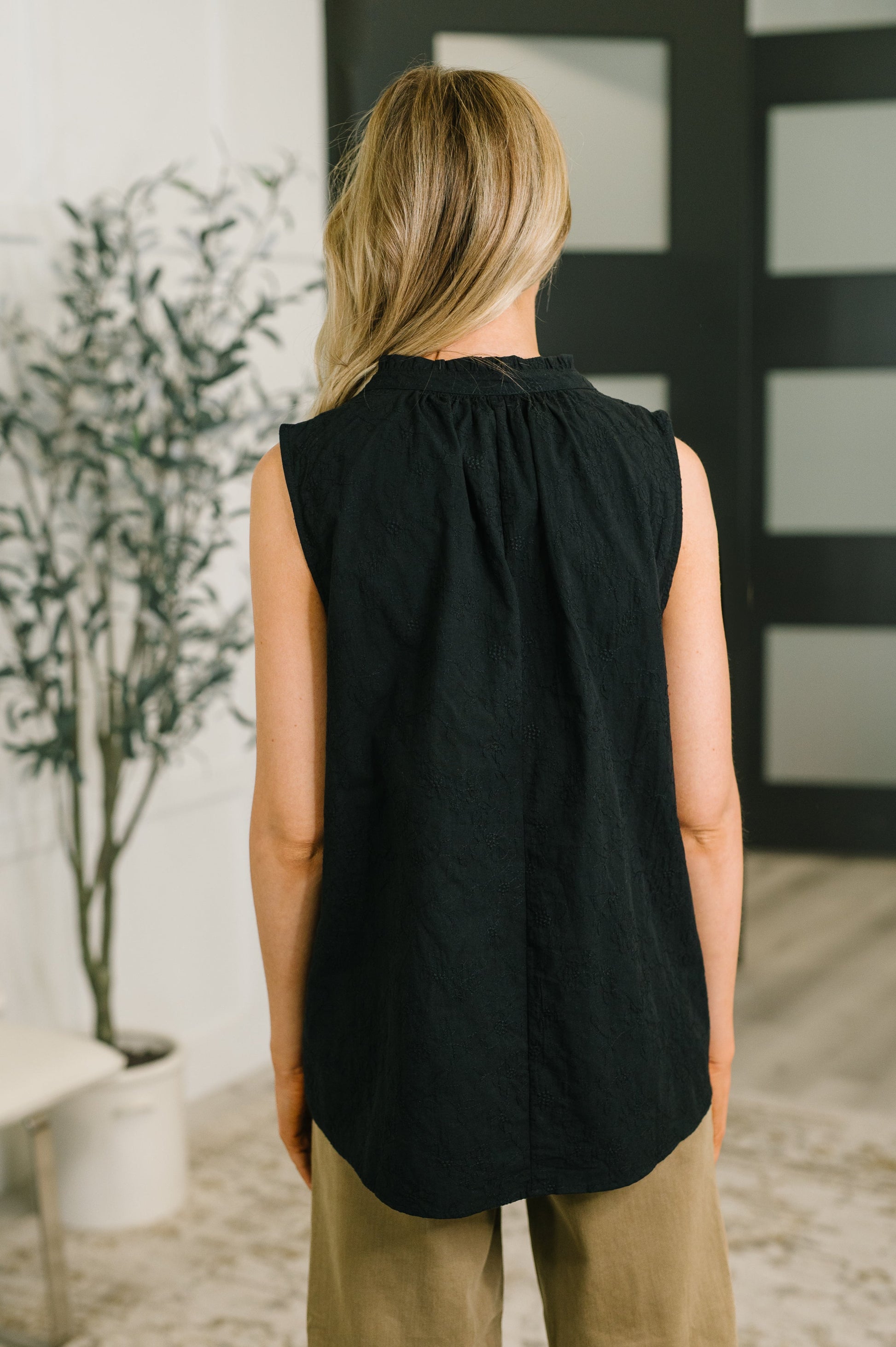 A woman viewed from behind wearing a sleeveless black top with a gathered neckline and beige pants, standing in an indoor room with a plant and dark paneled door in the background.