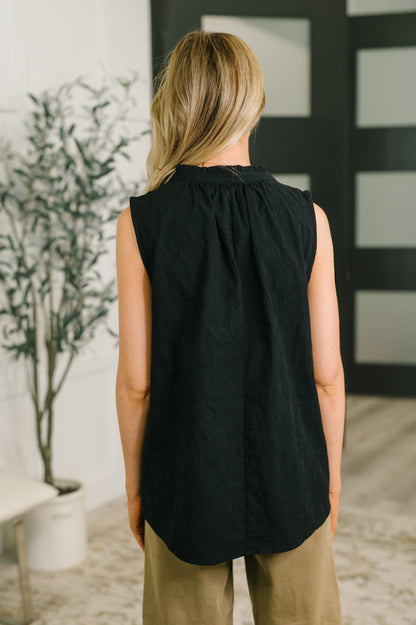 A woman viewed from behind wearing a sleeveless black top with a gathered neckline and beige pants, standing in an indoor room with a plant and dark paneled door in the background.
