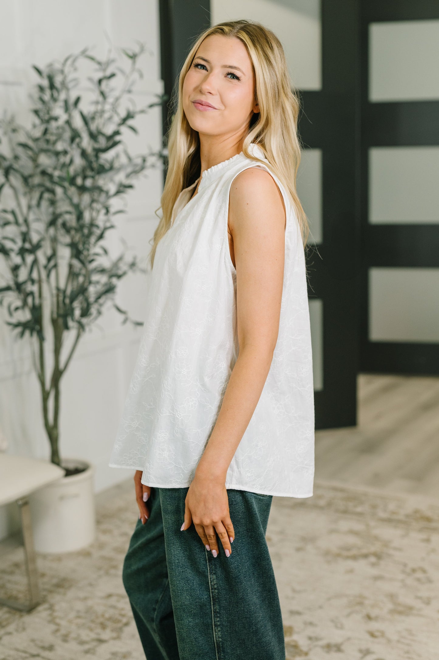 A blonde woman wearing a sleeveless white top and dark green wide-leg jeans, standing in a modern indoor setting with a potted plant and a black-framed glass door in the background.