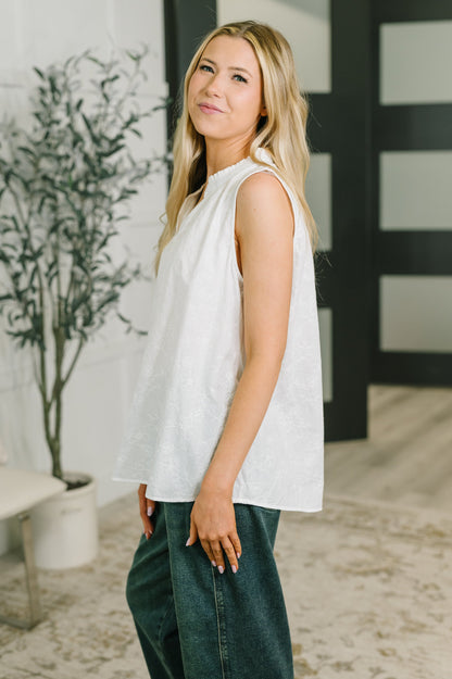 A blonde woman wearing a sleeveless white top and dark green wide-leg jeans, standing in a modern indoor setting with a potted plant and a black-framed glass door in the background.