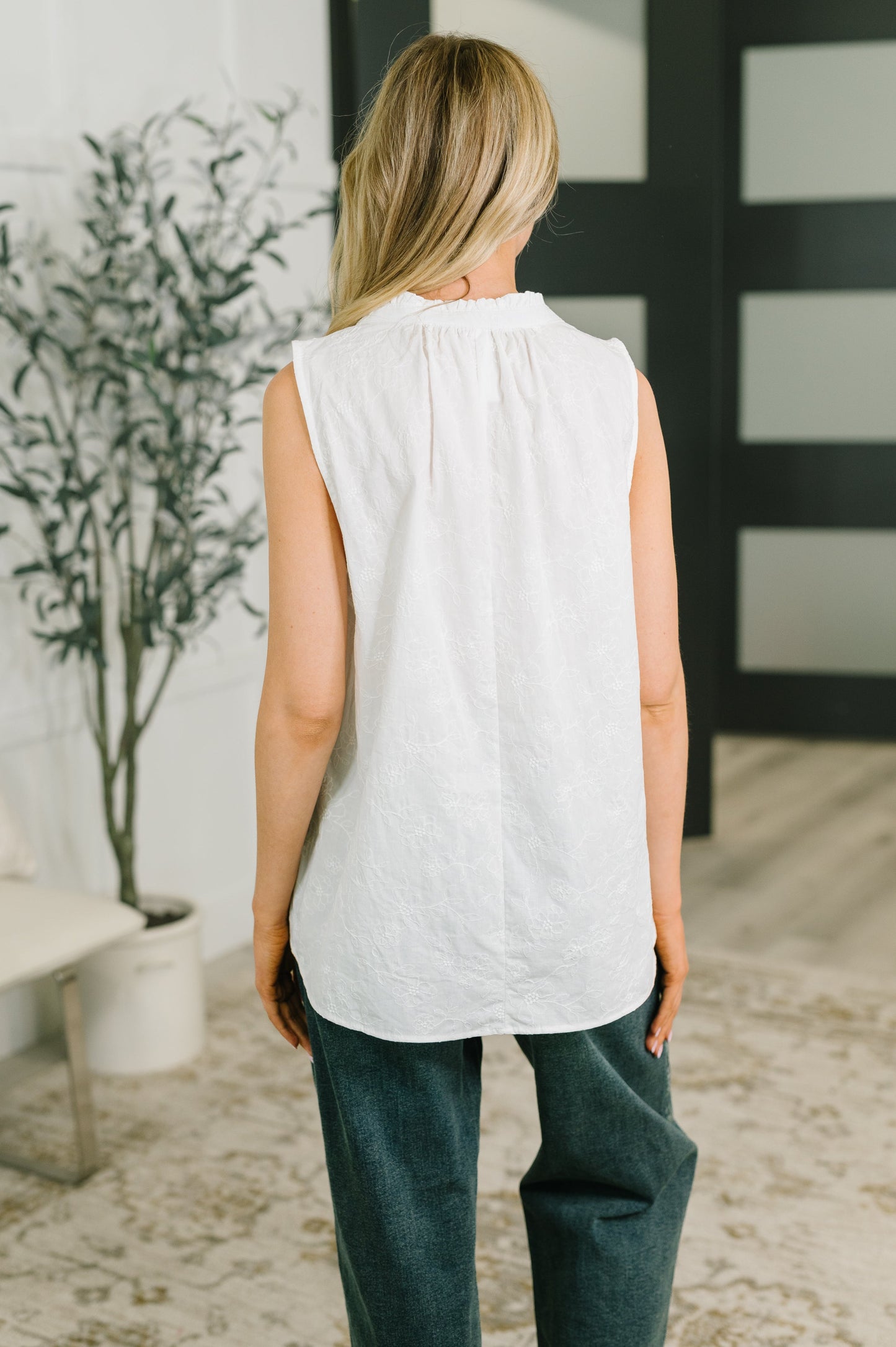 A woman viewed from behind wearing a sleeveless white top with a gathered neckline and dark green wide-leg pants, standing in a modern indoor room with a plant and black-framed window panels.