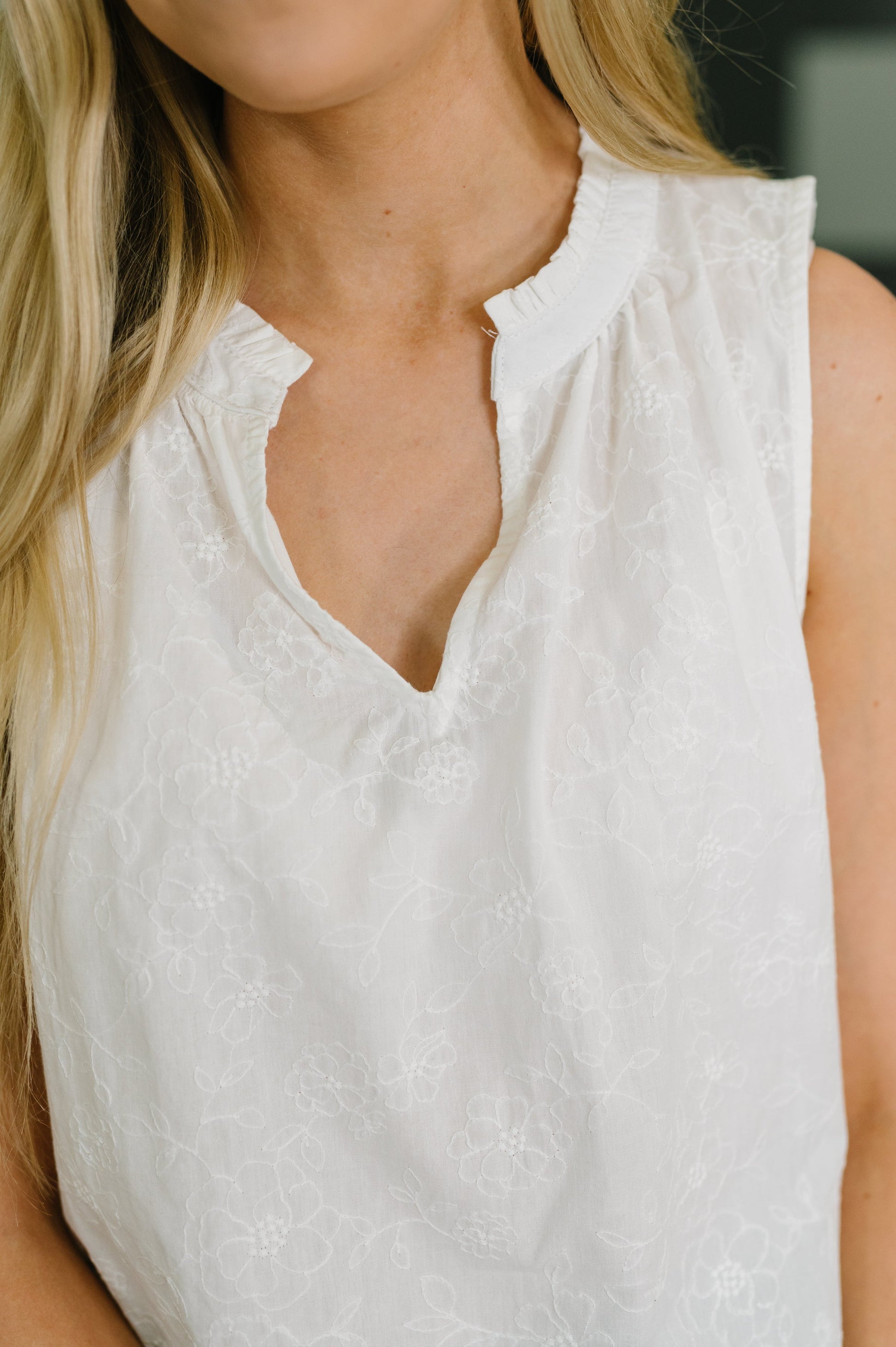 Close-up of a woman wearing a white sleeveless top with a textured, tonal floral lace-like pattern and a split neckline.