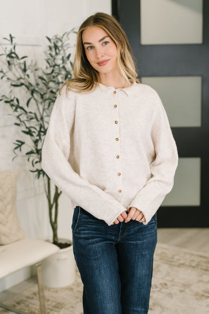 A woman wearing a cream-colored, long-sleeve knit button-up sweater and dark blue jeans, standing in a modern indoor setting with a plant and a bench in the background.