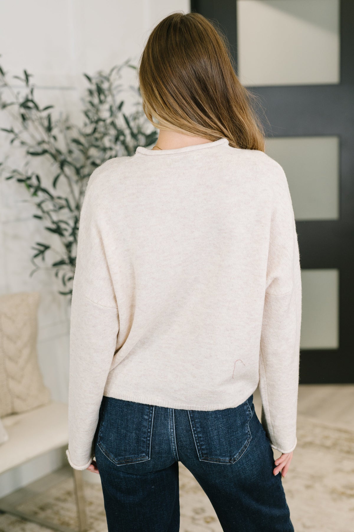 A woman viewed from behind wearing a light cream knit sweater and dark blue jeans in a modern indoor setting.