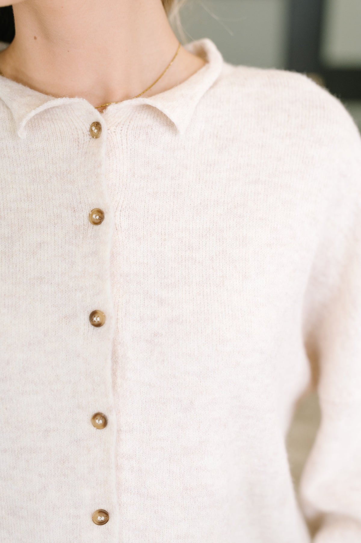 Close-up of a person wearing a cream knit cardigan with a collar and brown buttons, paired with a delicate gold necklace.