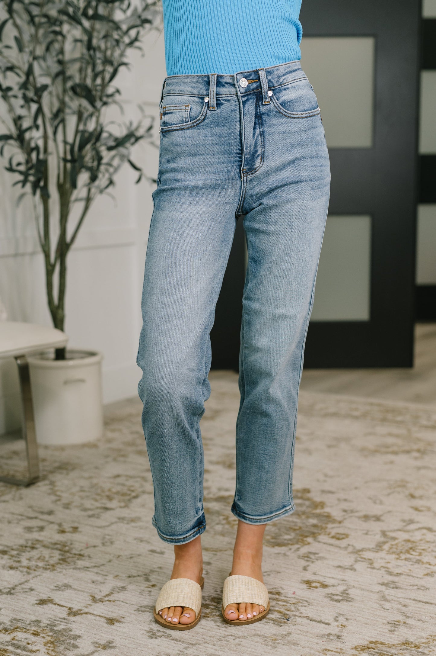 A person wearing light-wash blue cropped jeans with rolled hems, paired with a bright blue ribbed top and beige slide sandals, standing on a patterned rug in an indoor setting.