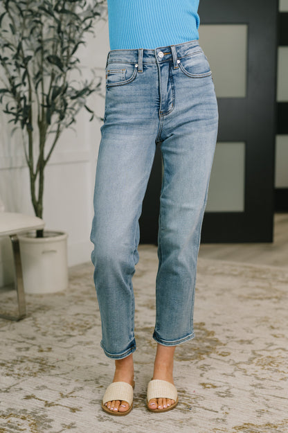 A person wearing light-wash blue cropped jeans with rolled hems, paired with a bright blue ribbed top and beige slide sandals, standing on a patterned rug in an indoor setting.
